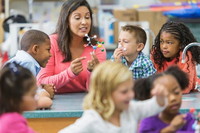 A teacher is holding a model of a molecule and talking while curious elementary children watch and listen.